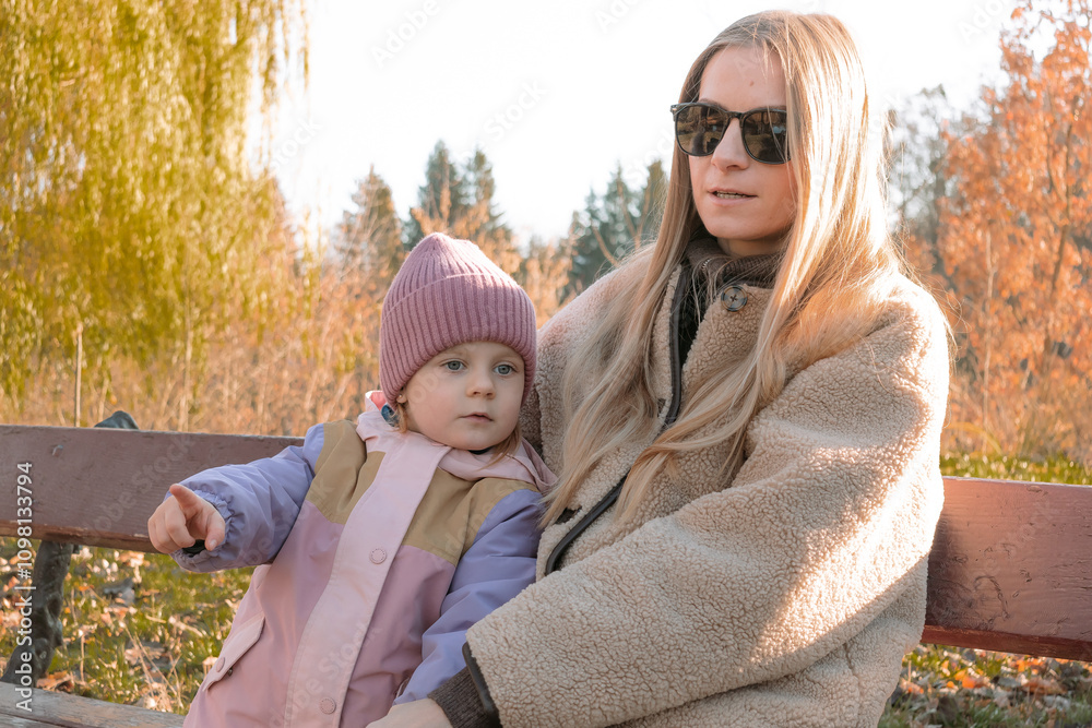 Fototapeta premium A happy girl and a young mother are sitting on a bench in autumn on a walk in nature