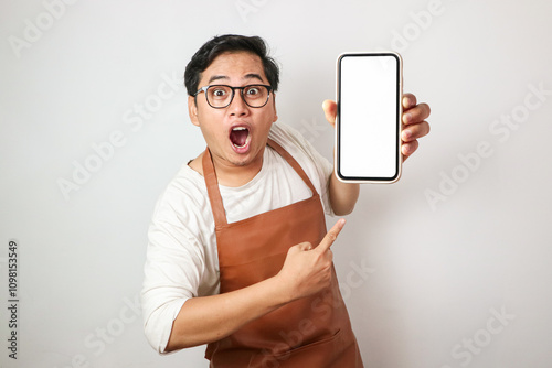 Surprised young Asian man barista barman employee wearing brown apron work in coffee shop using mobile phone isolated on white background. Small business startup concept