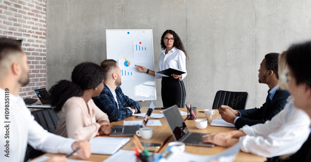 Fototapeta premium Latin Businesswoman Making Business Presentation Giving Speech For Colleagues Standing And Pointing At Blackboard With Charts And Graphs In Modern Office. Panorama, Selective Focus
