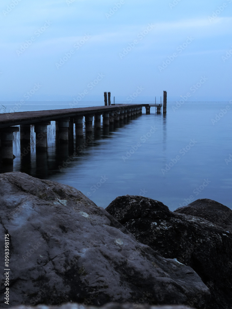 Obraz premium Muelle de madera en la costa de un lago con rocas, en un día nublado