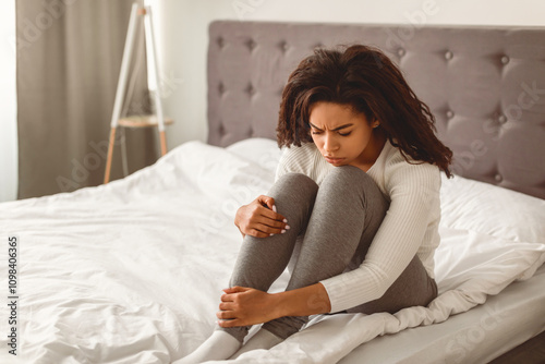 Depression Concept. Portrait of unhappy young African American woman sitting on bed alone and feeeling upset after breakup with her boylfriend or husband, hugging legs. Sad woman feeling lonely photo