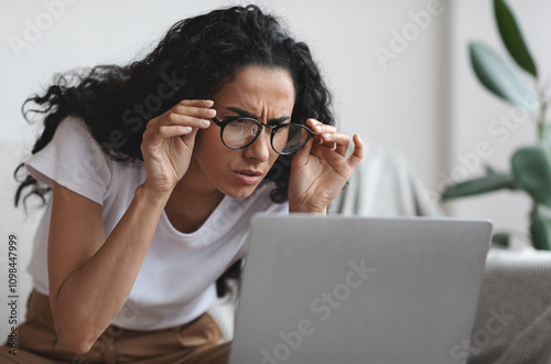 Young woman freelancer with bad eyesight using laptop, trying to work from home, copy space. Curly lady holding her glasses and squinting, looking at laptop screen, having vision troubles
