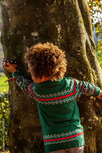 A toddler playing hide and seek behind a tree trunk. Bright and warm colours with golden sunlight. Multi ethnic boy.   