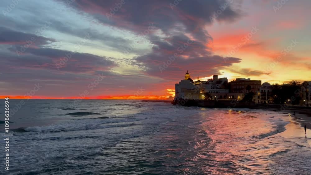 Video del atardecer pueblo Sitges, Barcelona. La luz del sol ha teñido las nubes de naranja creando reflejos en las aguas del mar y en la arena de la orilla, una estampa inolvidable, fondo de pantalla