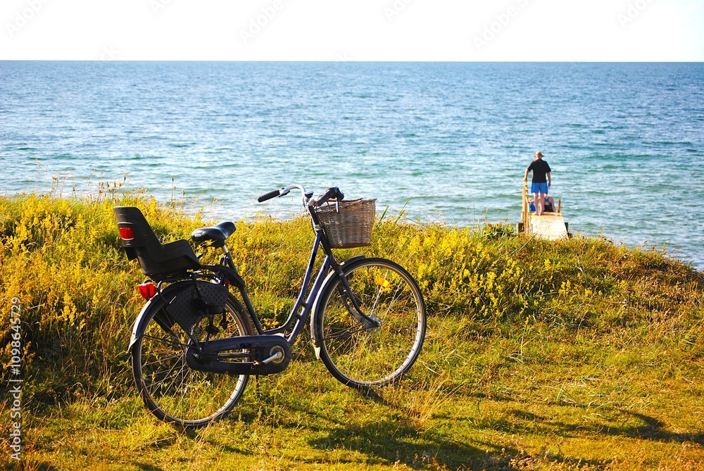 Fototapeta premium Man on pier with bicycle and grass in foreground with sea and clear sky. Denmark in summer.