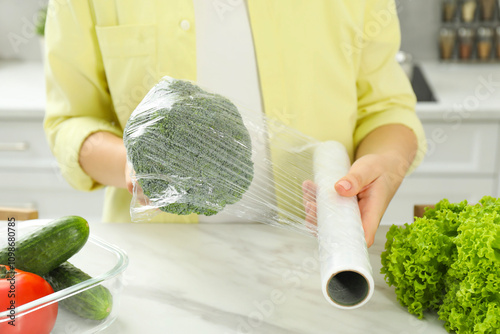 Woman putting plastic food wrap over broccoli at countertop in kitchen, closeup