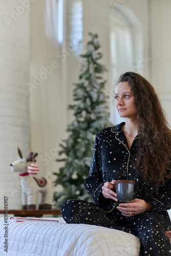 Cute young woman sits in bed in warm pajamas and holding mug - Christmas photo