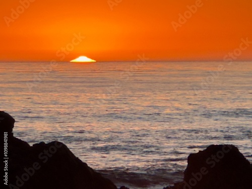 The image captures a stunning sunset over the Pacific Ocean near Cambria, California. The sun is partially set, casting a vibrant orange glow across the sky and reflecting on the calm ocean waters. photo