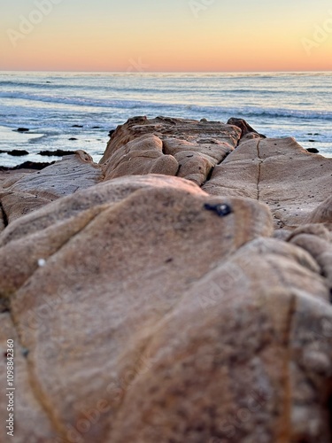 A stunning view of the rocky shoreline near Cambria, California, captured at sunset. The smooth, weathered rocks lead the eye towards the calm ocean waves and the vibrant, colorful sky.