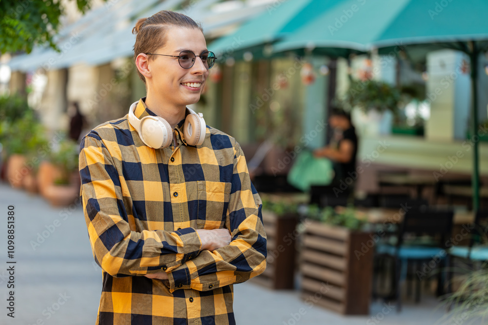 Obraz premium Portrait of happy man with headphones around neck smiling friendly, glad expression looking at side dreaming, resting, relaxation feel satisfied good news outdoors. Young guy in urban city street.