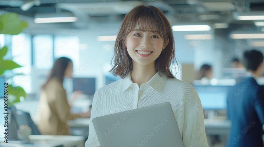 Obraz premium Smiling Japanese businesswoman standing in an office, holding her laptop. She is wearing casual clothes, and other people working behind her are blurred.,generated ai 