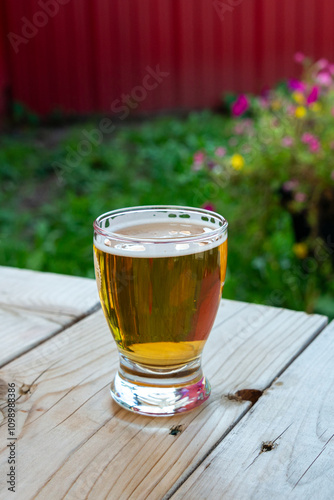 A side view of a small sample glass of pale ale craft beer. The liquid has a yellow tint. A clear beer glass sits at the edge of a wooden table at a microbrewery. The background has pink flowers. photo