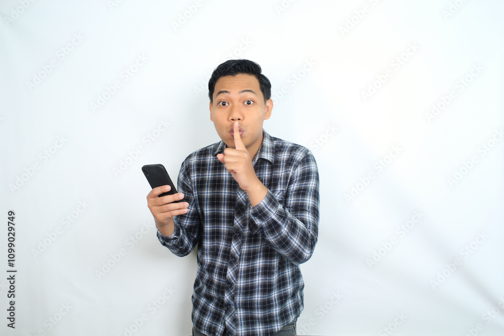 Fototapeta premium asian man holding mobile phone showing silent gesture with finger on lips isolated on white