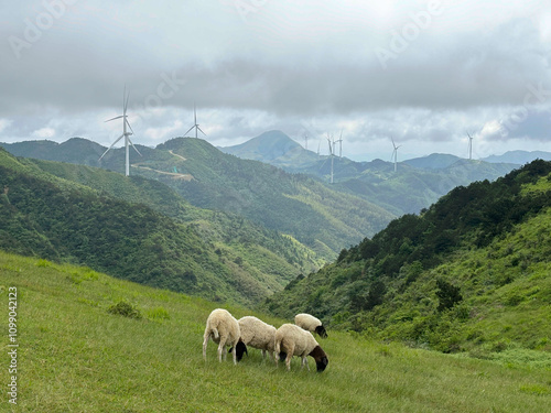 A flock of sheep wanders around the mountains with windmills in the background.