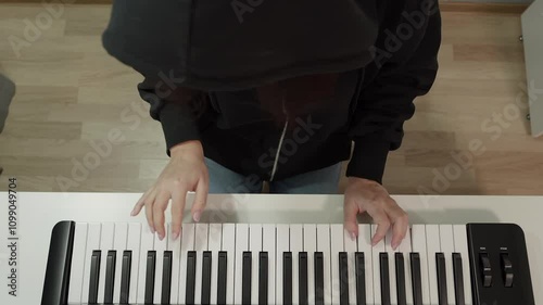 High angle view of musician playing a small synthesizer keyboard wearing a black hooded sweatshirt while sitting at white table in home studio