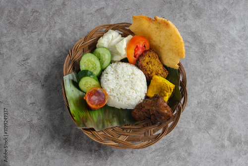 Delicious plate of Indonesian food featuring ayam bakar (roasted chicken), served with a mound of white rice, crispy cracker, tempeh, and fresh vegetables like cucumber and tomato on a woven basket photo