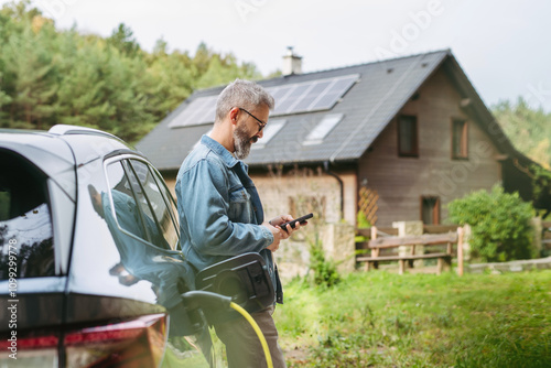 Man charging electric car in front of his house, plugging the charger into the charging port. House with solar panel system on roof behind him. photo