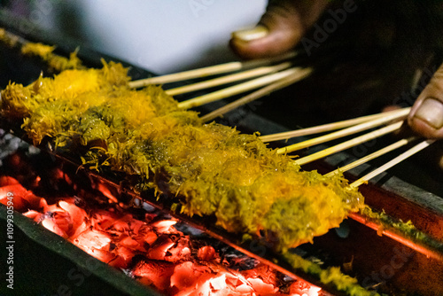 Close up of Sate kelapa or sate klopo or coconut satay, chicken or meat satay with coconut ingredient on red fire grilling by people. Traditional satay from Surabaya, East Java, Indonesia. photo