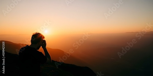 A silhouette of a person sitting on a mountain peak at sunset, with the sun peeking through the hazy sky and mountains in the background