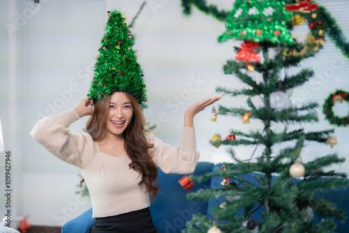 Happy young woman wearing a christmas tree hat is showing a decorated christmas tree with her hand while smiling and touching her hat photo