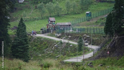 A shot of SUV vehicle or car moving though a mountainous scenic landscape surrounded by mountains and green meadow an pine tree forest during rainy or monsoon season. 
