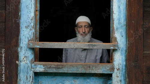 An aged or elderly Muslim Indian Kashmiri man in ethnic attire sitting indoors, looking outside from the frame of the window of an old village wooden house in a Himalayan mountainous region of Kashmir