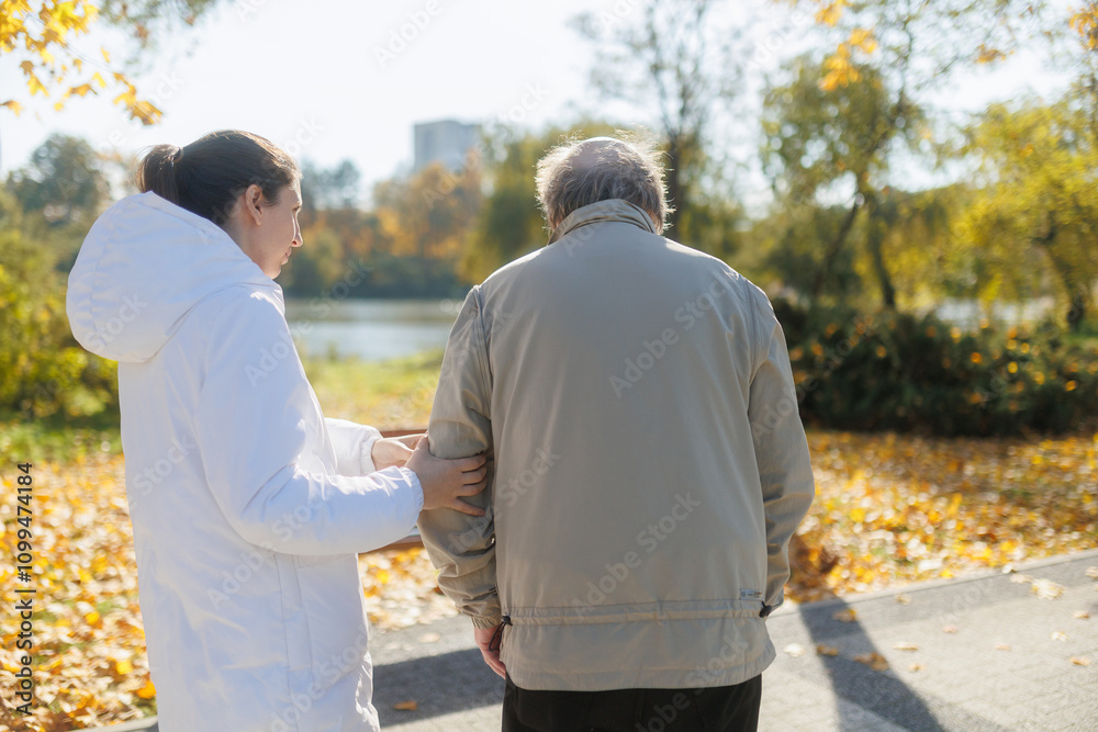 Obraz premium Elderly man and nurse in autumn park, back view of outdoor companionship