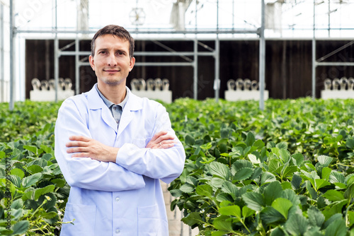 A confident man in a lab coat stands in a greenhouse, surrounded by lush green plants, showcasing agricultural expertise and innovation. photo