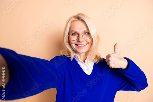 Confident mature woman in blue sweater gives thumbs-up against a beige background photo