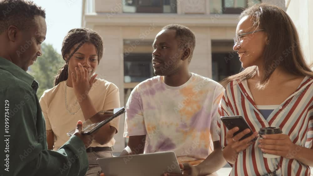 Three young students sit on break chatting outdoors. Female and male coworkers gather and brainstorm together near urban building using gadget. Young man and woman communicate discussing and talking