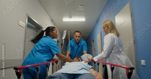 Medical workers carry a patient on a gurney along the hospital corridor. A woman holds an oxygen mask near his face. They're running