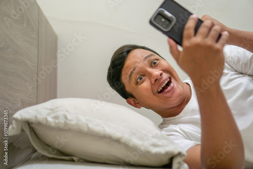 Young Asian man lying on bed while using smart phone with shocked expression. Man shows surprised expression while holding smartphone in bed at night. photo