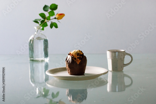 A cupcake on a white plate and a glass vase with a yellow rose, a small beige cup for tea or coffee. Still life with food and drink. Chocolate maffin or muffin - traditional American dessert.  photo