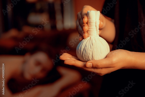 Hot herbal ball spa massage body treatment with masseur's hand hold or show herb bag at spa. Tranquil and serenity of aromatherapy recreation in warm lighting of candles at spa salon. Quiescent