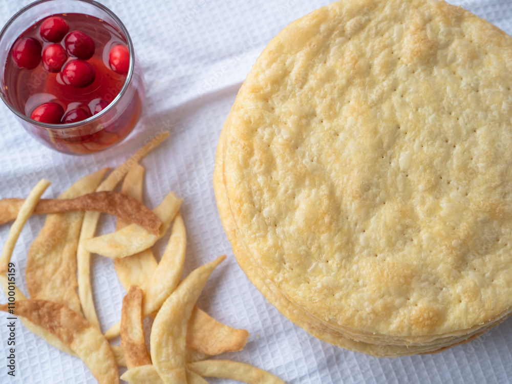 Top view over a pile of baked thin round buttery layers, dough scraps on a white waffle towel ready to make a shortcrust puff pastry Napoleon cake. A glass of red berry drink nearby.