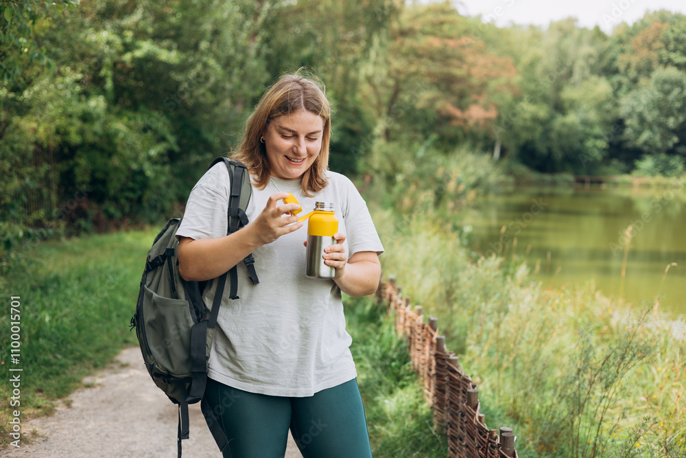 Happy smiling 30s women with backpack is holding thermos mug. Hot tea or other beverage on summer day. Lifestyle adventure concept. Woman drink cold water from the bottle