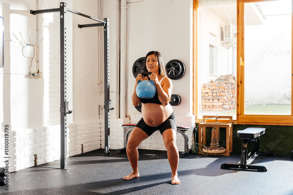Fototapeta premium Pregnant Chinese Woman Performing Kettlebell Squats in a Bright Home Gym During Morning Exercise Routine