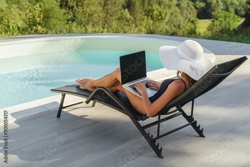 A woman in a white sunhat relaxes on a lounge chair by the pool, working on her laptop. Balance of remote work, leisure, and relaxation in an outdoor environment. Space for text