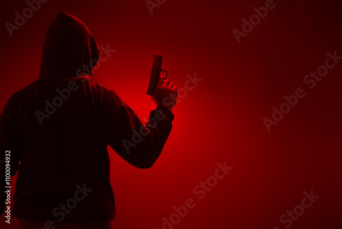 An unidentified man in a black hood and black mask stands holding a gun in various poses. Light is shining from above. Looks scary. Isolated over film studio background. Armed person.