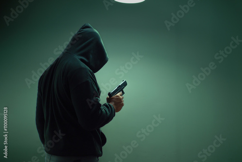 An unidentified man in a black hood and black mask stands holding a gun in various poses. Light is shining from above. Looks scary. Isolated over film studio background. Armed person.