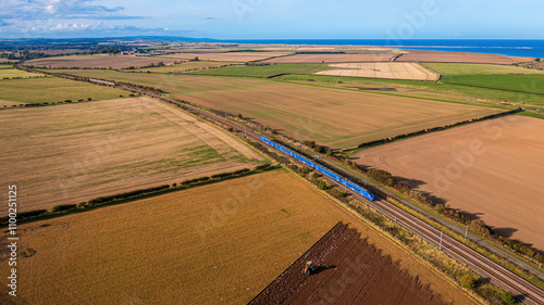 Aerial view of expansive farmland and train tracks alongside the coastline under a blue sky with clouds, Fenham, United Kingdom. photo