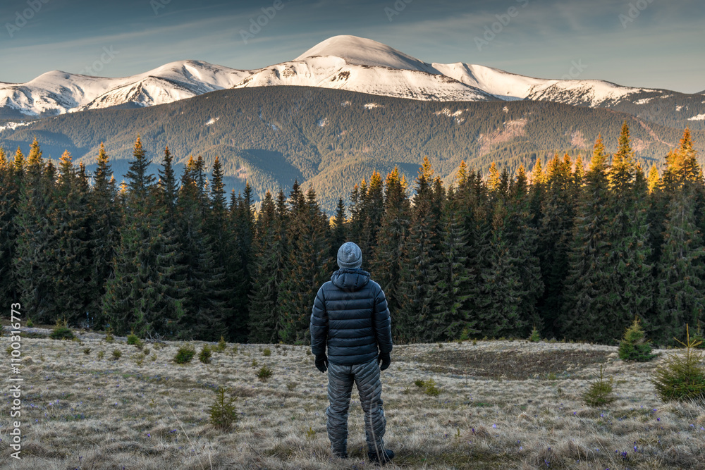 Obraz premium Rear view of young man in warm clothing embarks on a hiking trip at sunrise, surrounded by the breathtaking backdrop of snowy mountains