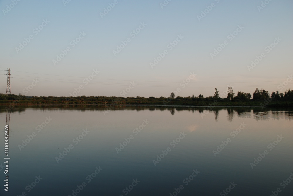 Naklejka premium Windless evening on the lake. Summer evening clear light blue sky without clouds. Smooth flat surface of water in forest lake. Sky and plants growing on opposite shore of lake are reflected from water