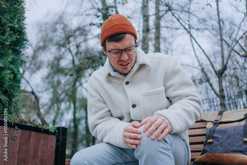 Young man wearing glasses and winter clothes is suffering from knee pain, sitting on a bench in a park, holding his leg with both hands photo