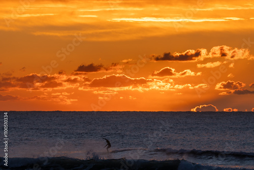 Surfista en acción durante un vibrante amanecer sobre el mar, con el sol descendiendo entre nubes doradas y olas suaves. photo