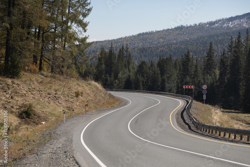 Long curved asphalt road running along the mountain pass. Crash barrier are along the road. Golden autumn roadscape with yellow trees and wild nature. Hilly taiga landscape in the distance photo