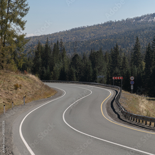 Long curved asphalt road running along the mountain pass. Crash barrier are along the road. Golden autumn roadscape with yellow trees and wild nature. Hilly taiga landscape in the distance