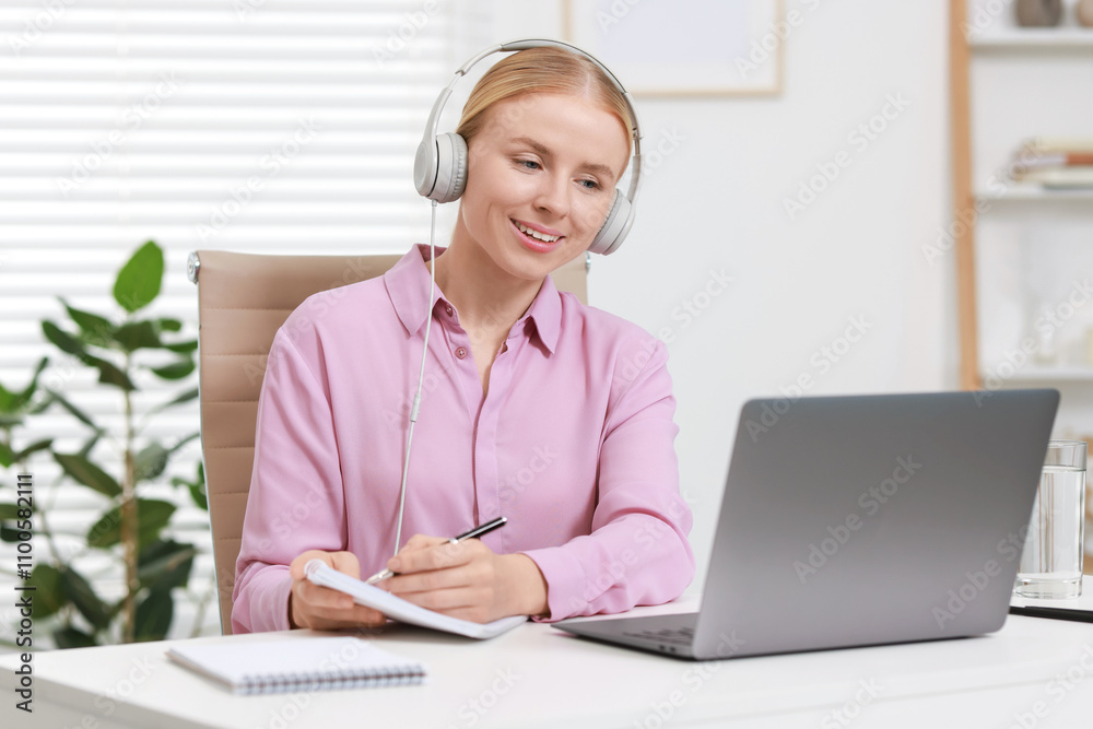 Interpreter in headphones taking notes while having video chat via laptop at table indoors Interpreter in headphones taking notes while having video chat via laptop at table indoors