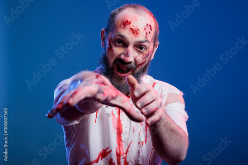 Portrait of walking dead corpse with bloody wounds isolated over blue studio background. Brain eating scary zombie growling creepily in front of camera, starving for human flesh photo