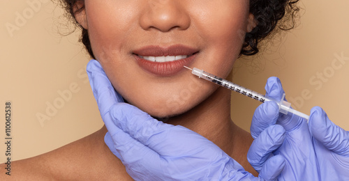 A woman is smiling while receiving a cosmetic injection for lip enhancement. The procedure takes place in a clinic with soft, neutral background colors, cropped photo